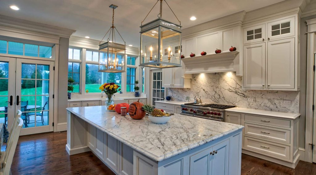 All-white kitchen designed in Brighton beaded inset cabinetry with oversize pendant lights above a large island and a slab backsplash behind a gas range.